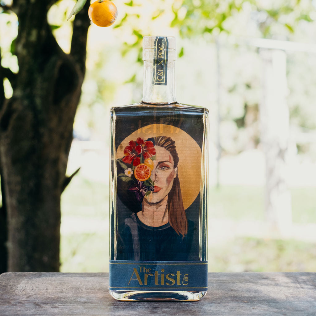 Bottle of 'Artist' gin with a portrait label, placed against a natural background with orange trees.