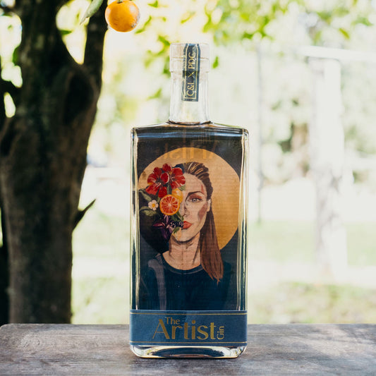 Bottle of 'Artist' gin with a portrait label, placed against a natural background with orange trees.